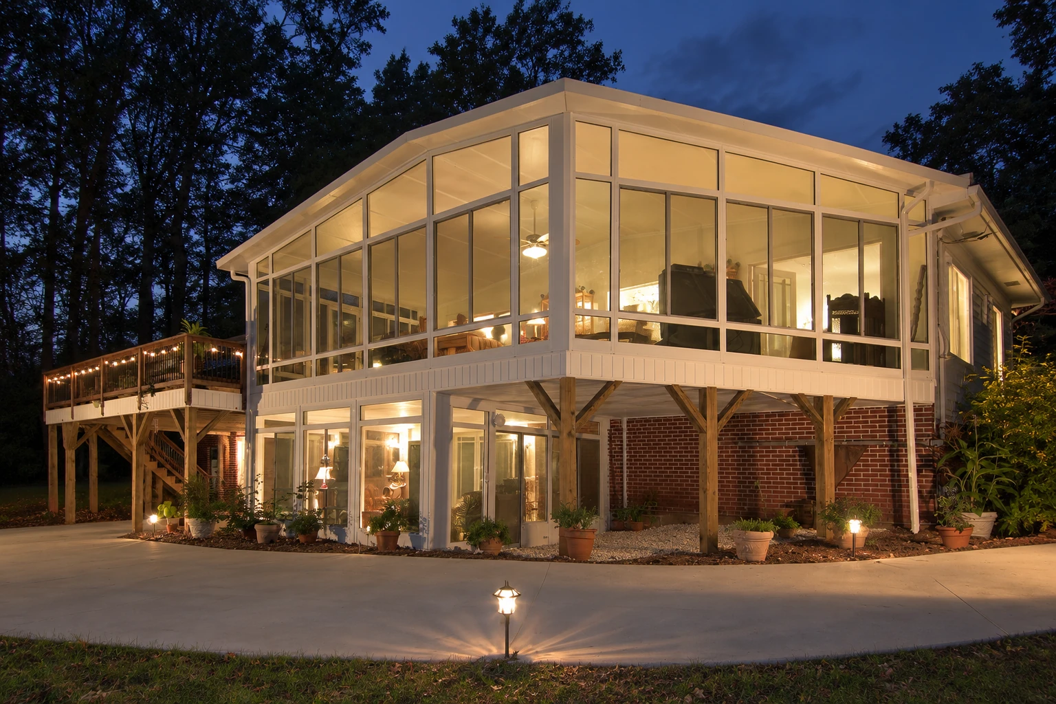 A raised, glass-walled sunroom glows at dusk beside a wooded yard, with a deck and lights along the walkway.