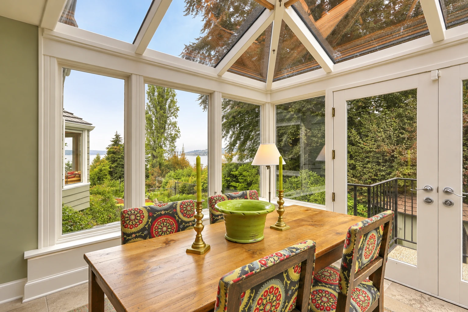 A solarium with a white-framed glass roof, a wooden dining table, and views of trees and a lake through wide windows.