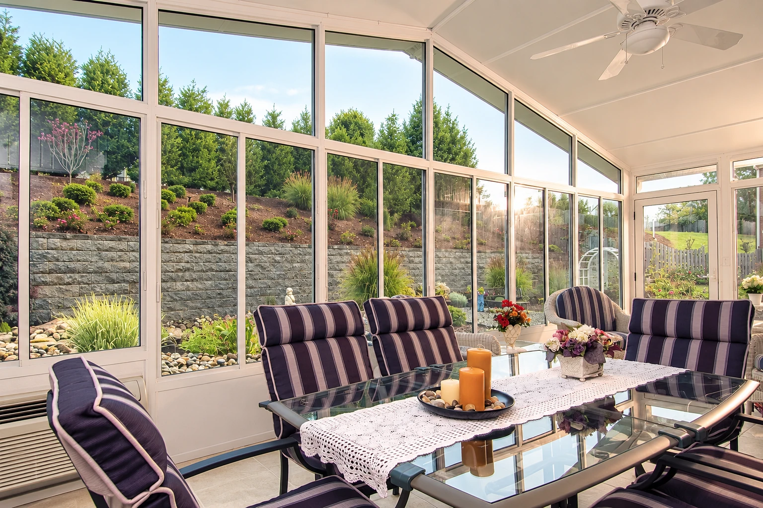 Sunroom with white-framed windows, striped lounge chairs and a glass table overlooking a landscaped garden.