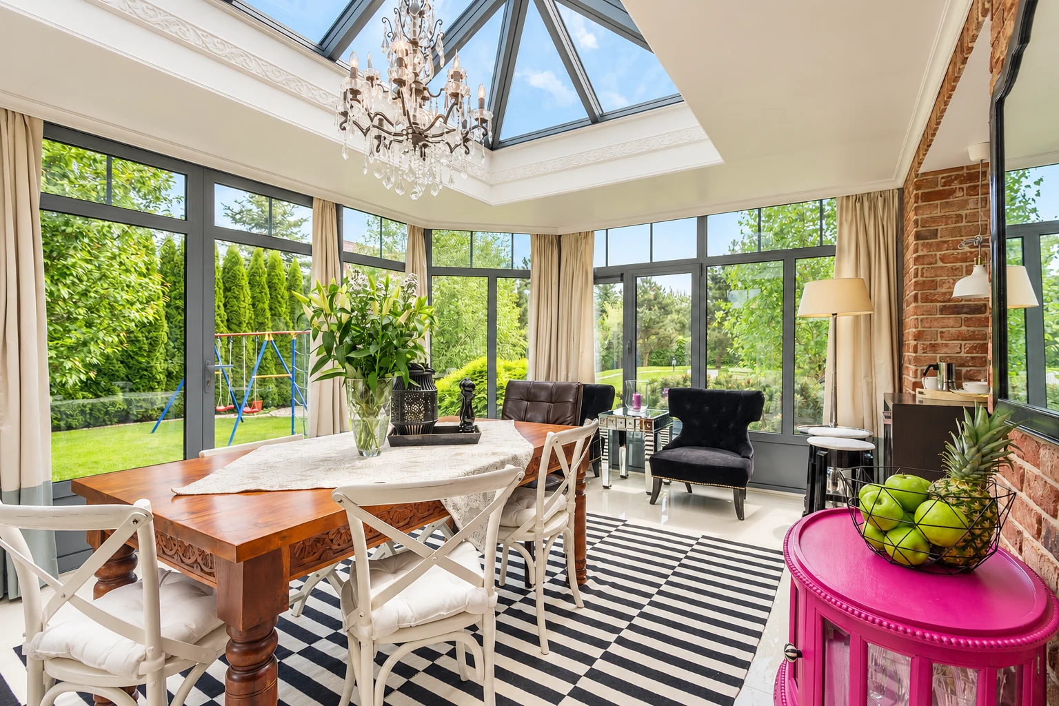 Bright sunroom with a glass-paneled roof, dark-framed windows, a dining table, and a black-and-white striped rug.